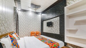 A hotel room with a bed, two chairs, a TV, and a shelf unit. - Fabexpress Milan Palace (Faizabad)