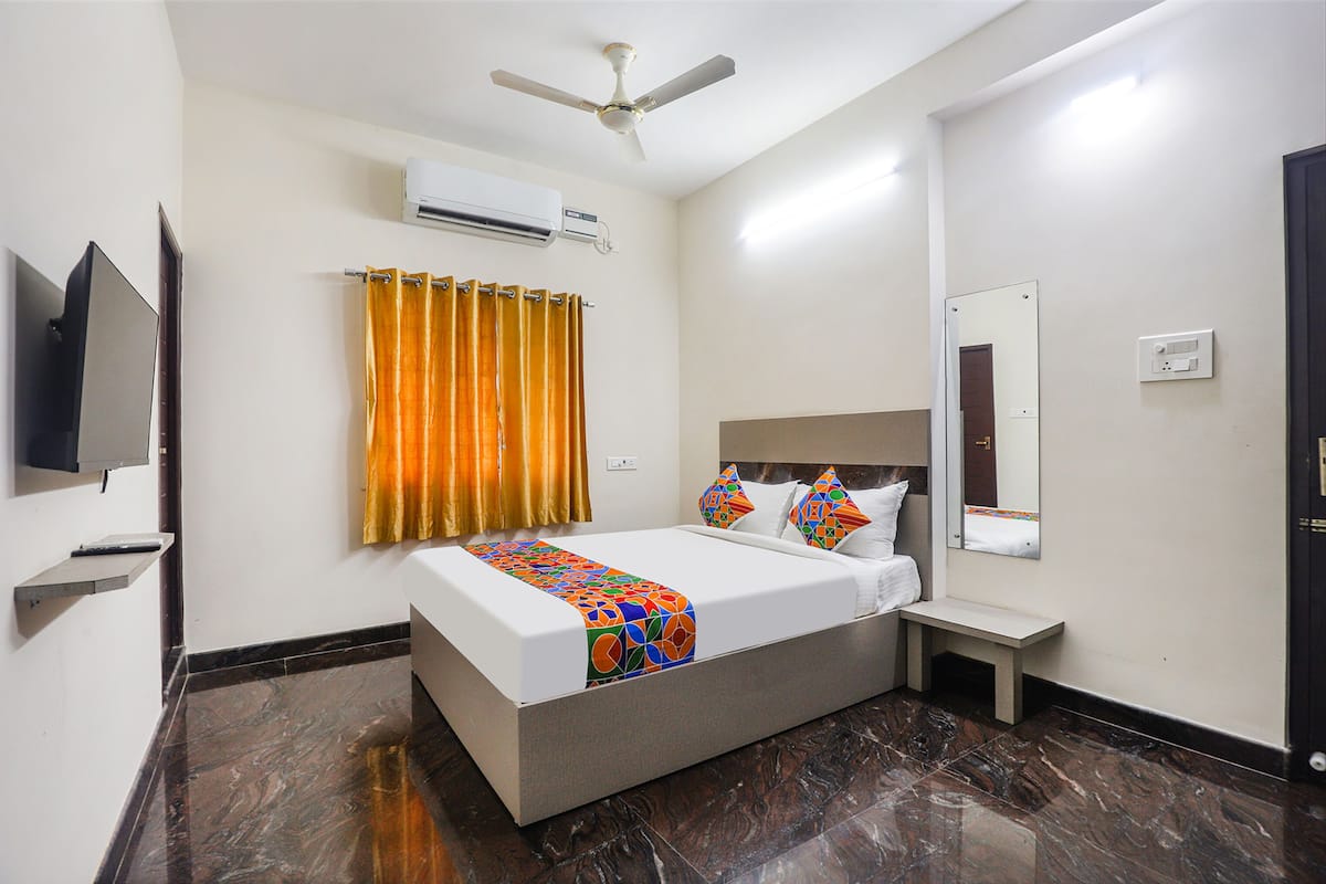 Habitación de hotel con cama, televisión, aire acondicionado, ventilador de techo, espejo y mesita de noche pequeña.
