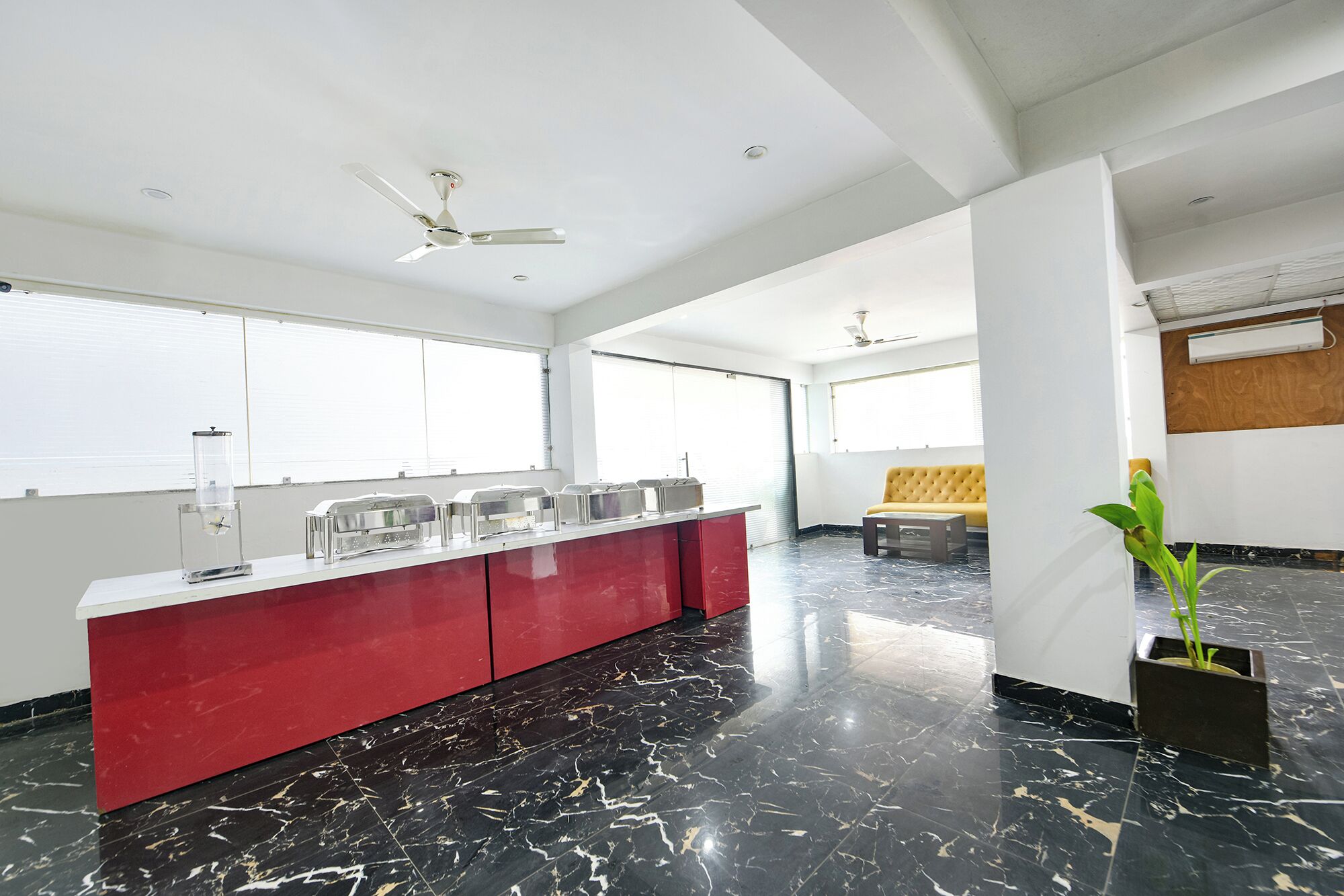 Um hall de hotel moderno com piso de mármore, balcão vermelho com recipientes de vidro, sofá amarelo e ventoinha no teto.