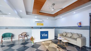 A hotel reception area with a check-in counter, a sofa, a chair, a small table, and a ceiling fan. - Fabhotel Vibe (Noida)