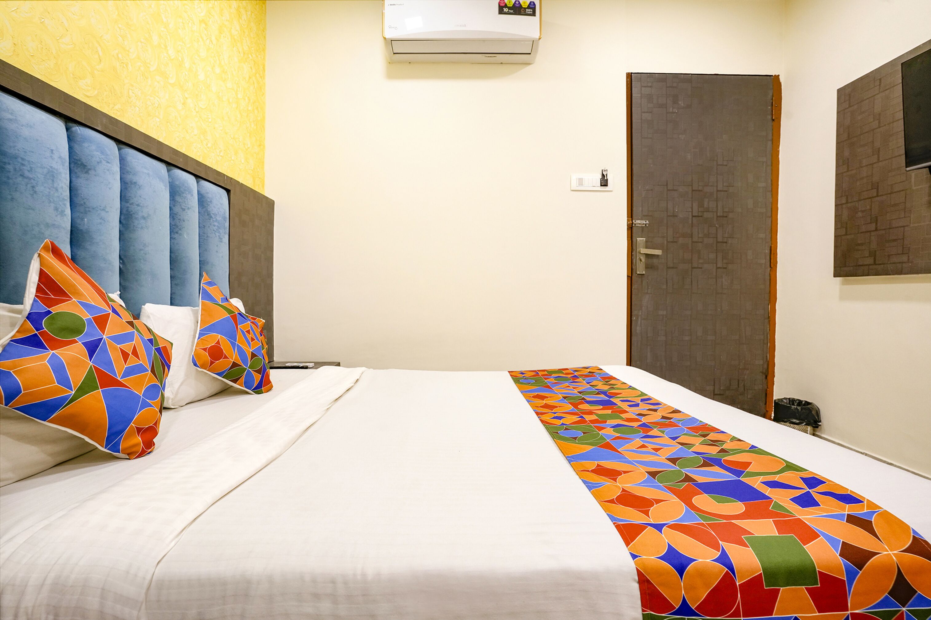A hotel room with a bed, colorful pillows, a patterned bed runner, a wall-mounted air conditioner, and a door.