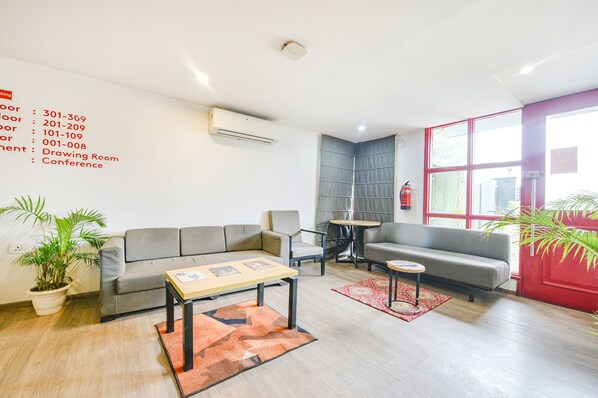 A modern living room with a grey sofa, a coffee table, and a small side table. There are potted plants and a red-framed window. - FabHotel Comfort Residency I - Nr. Artemis Hospital Gurgaon (Gurugram)