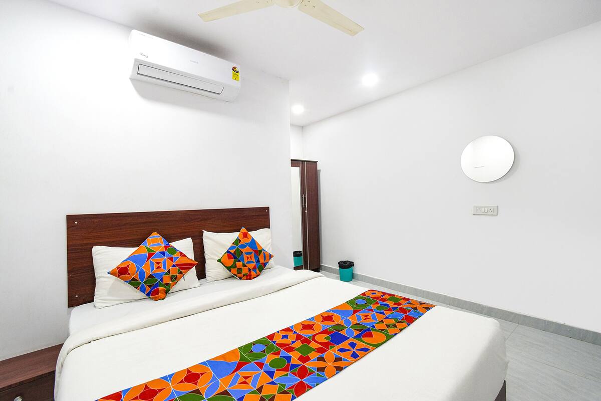 A modern hotel room with a bed, a ceiling fan, an air conditioning unit, a bedside table, a trash bin, and a round mirror on the wall.