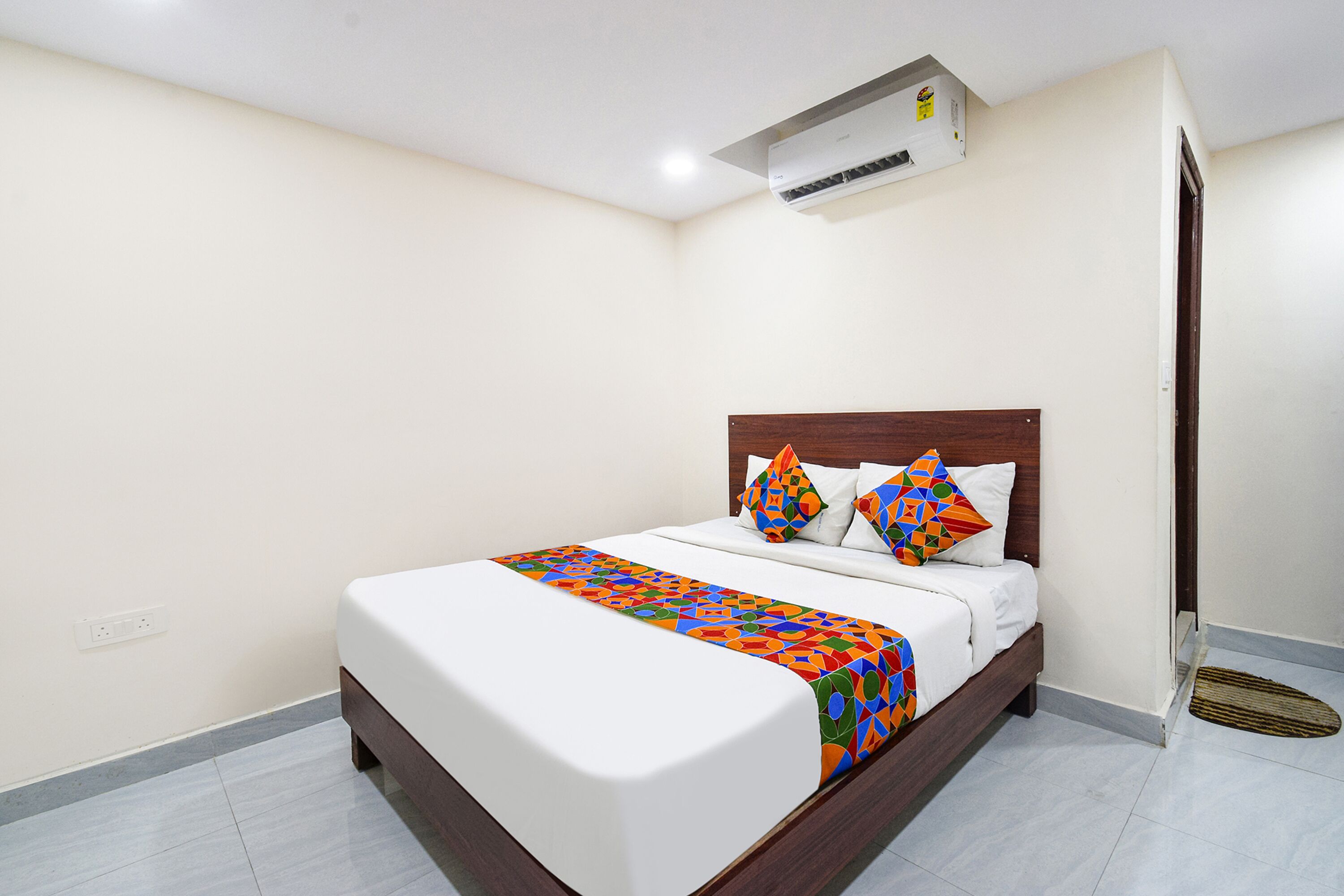Una cama bien hecha con un edredón de estampado geométrico y colores vivos, un cabecero de madera y una pared blanca con aire acondicionado instalado en el techo.