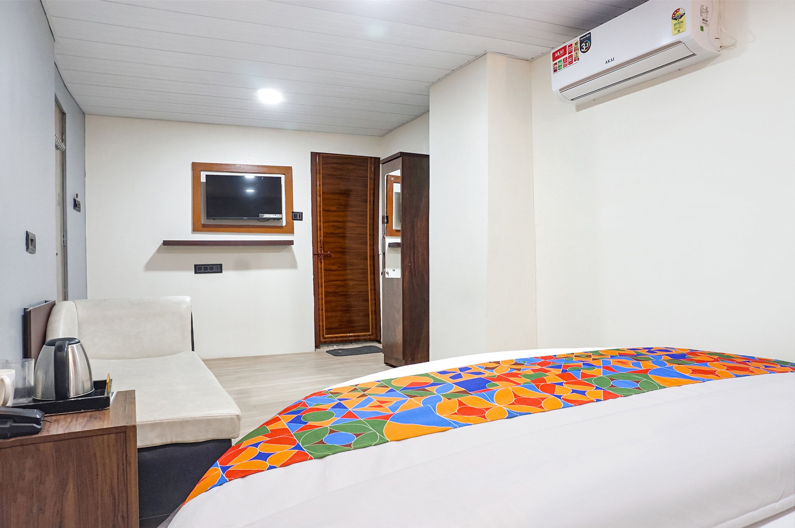 A hotel room with a bed, a desk with a lamp, a TV mounted on the wall, and an air conditioning unit.