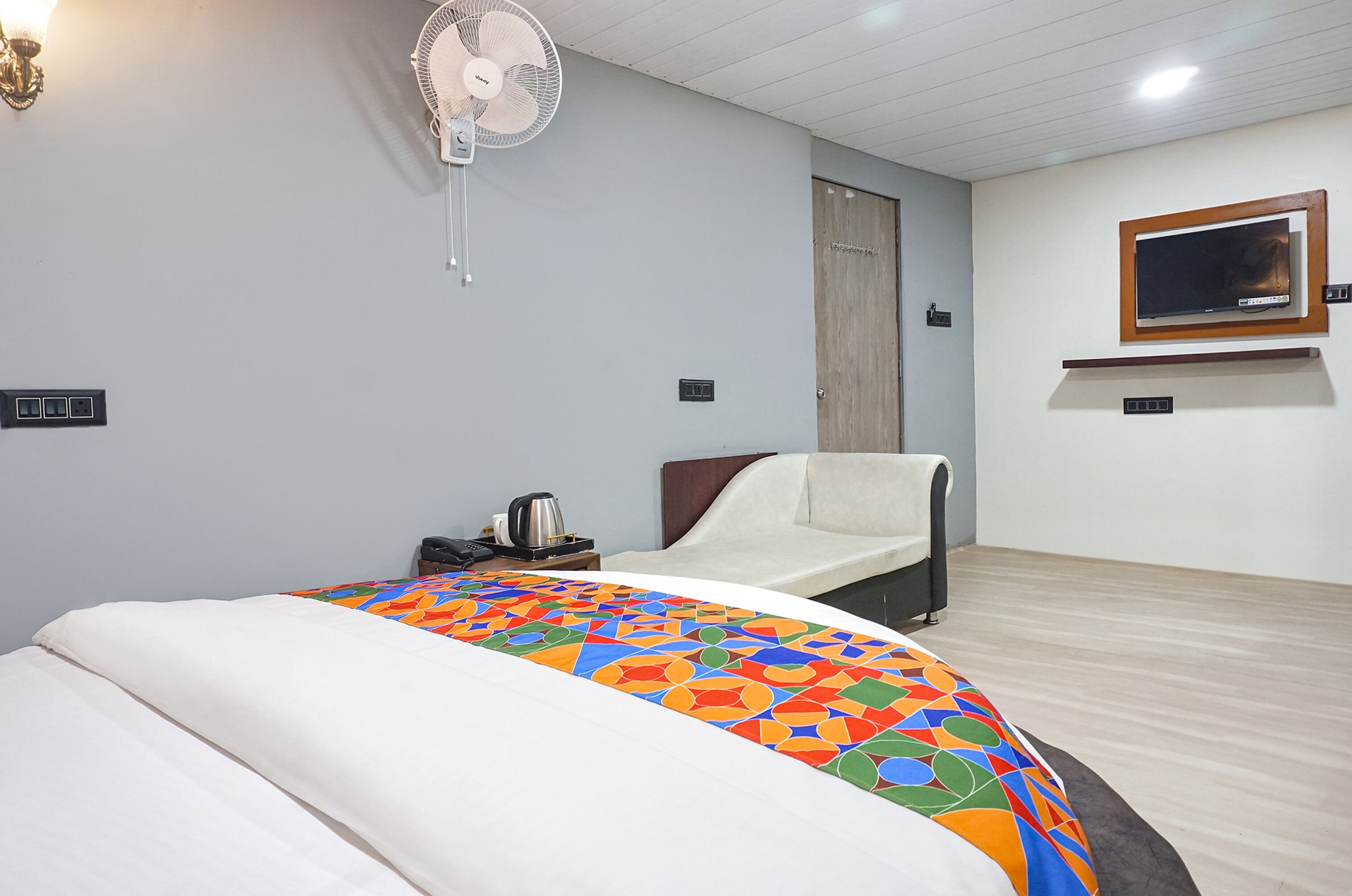 Habitación de hotel con cama, sillón, televisión montada en la pared y ventilador de techo.