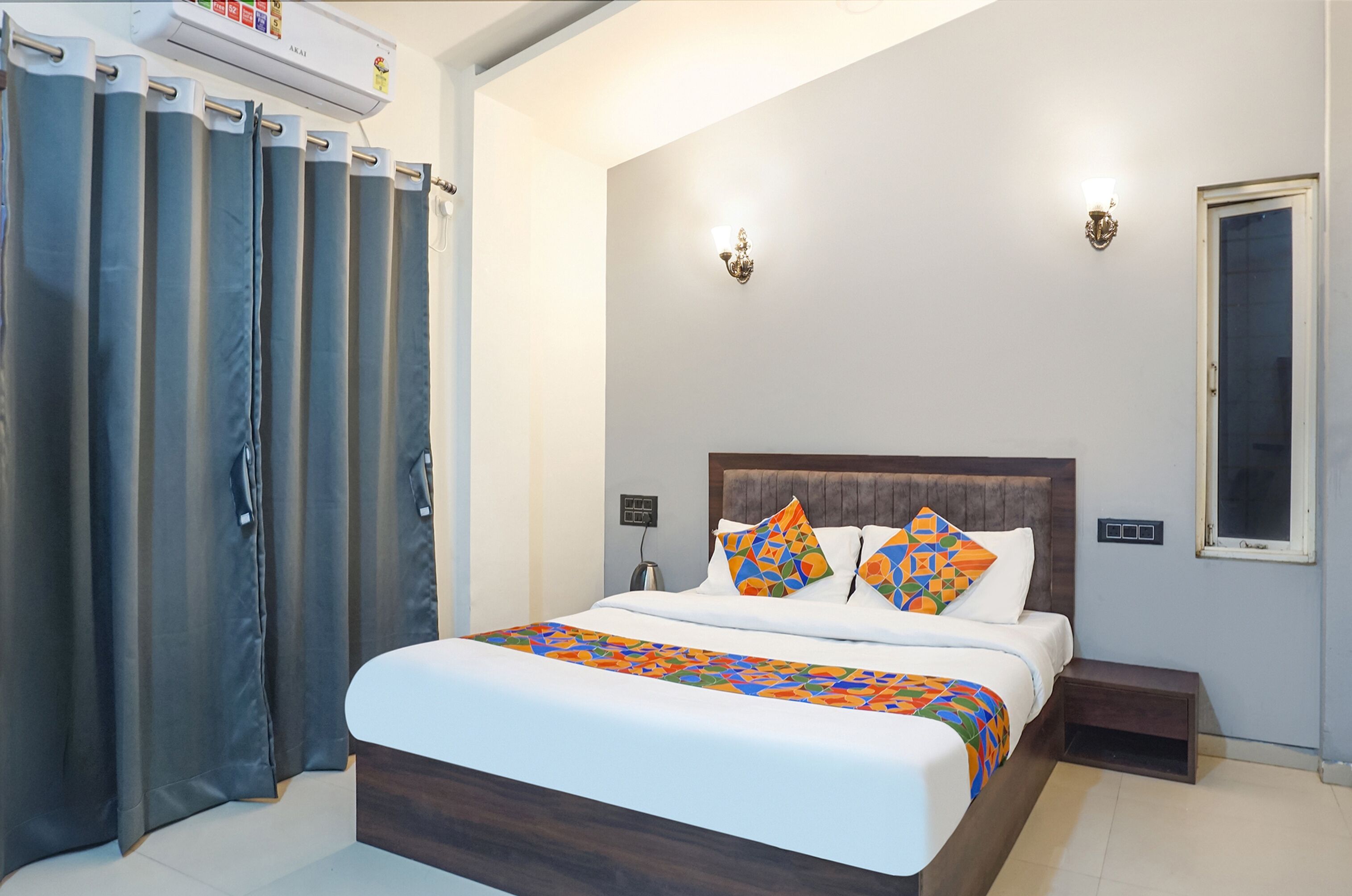 A hotel room with a large bed, a nightstand, a wall-mounted lamp, and a window with curtains.