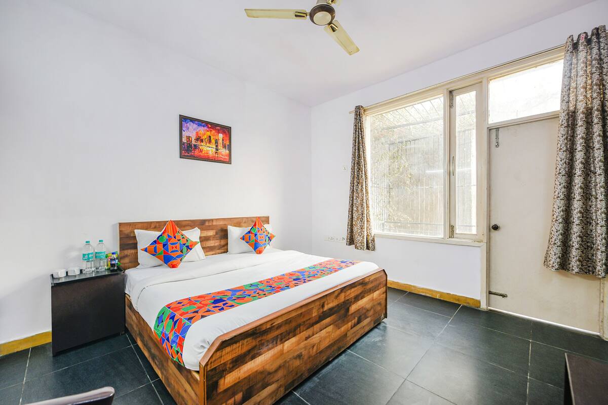 Une chambre à coucher avec un lit en bois, une table de chevet, un ventilateur de plafond, une fenêtre avec des rideaux et un tableau accroché au mur.