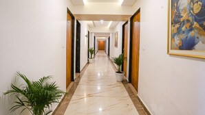 Un couloir d’hôtel avec un sol en marbre, des portes en bois, des plantes en pot et un tableau encadré accroché au mur.