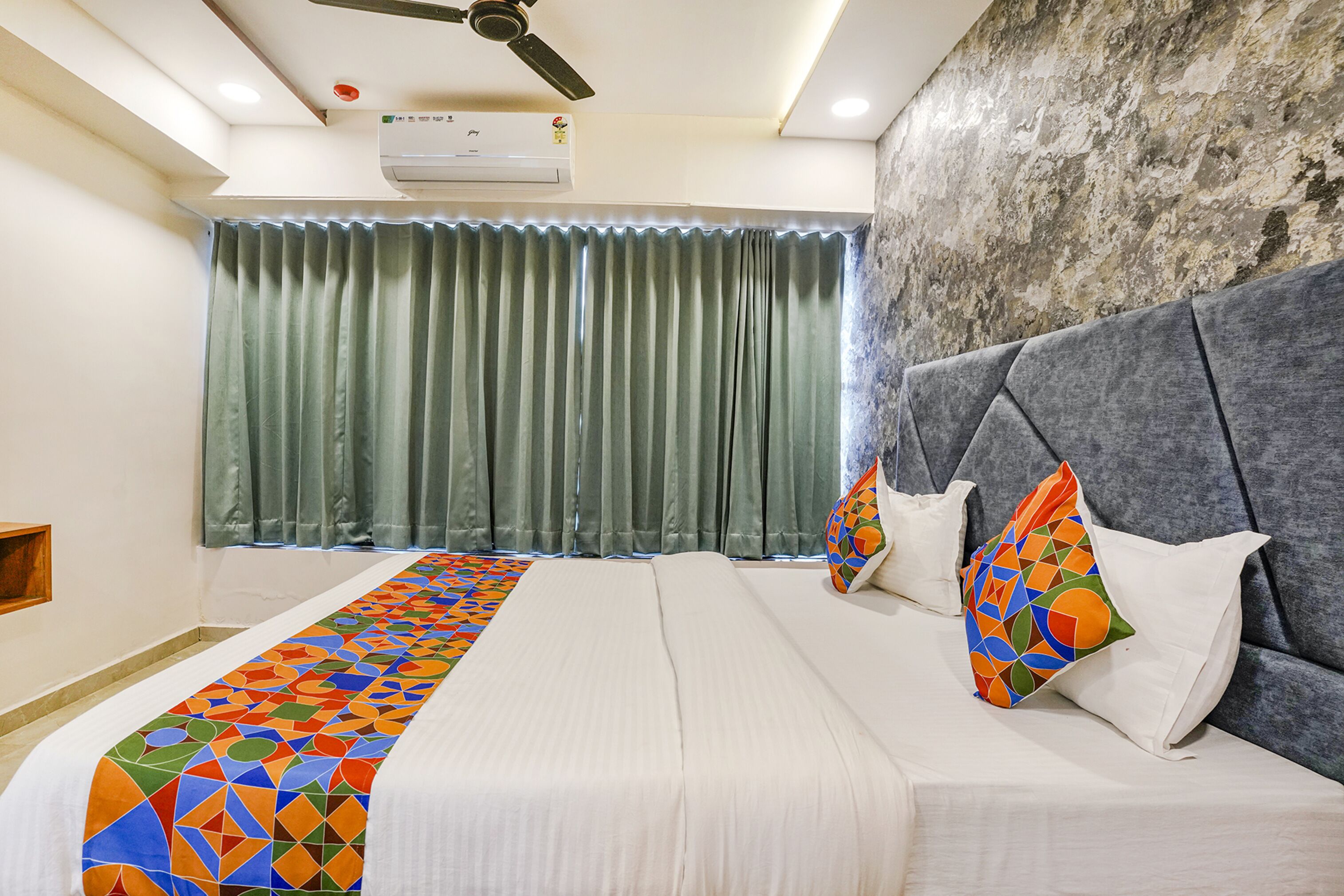 Una habitación de hotel con una cama grande, un cabecero tapizado en gris, un corredor de cama estampado y una cortina verde.