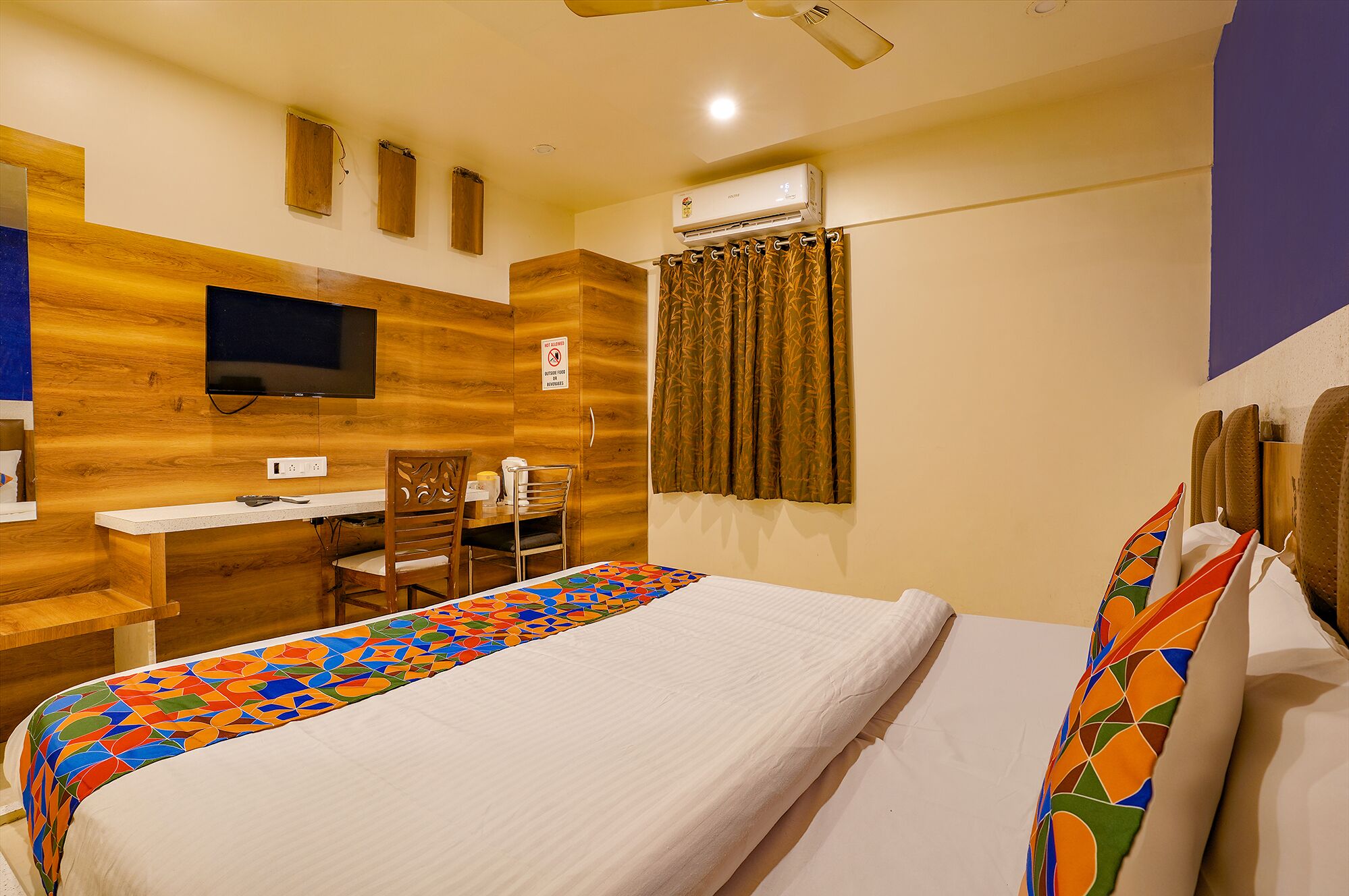 A hotel room with a bed, a desk, a chair, a television, and an air conditioning unit.