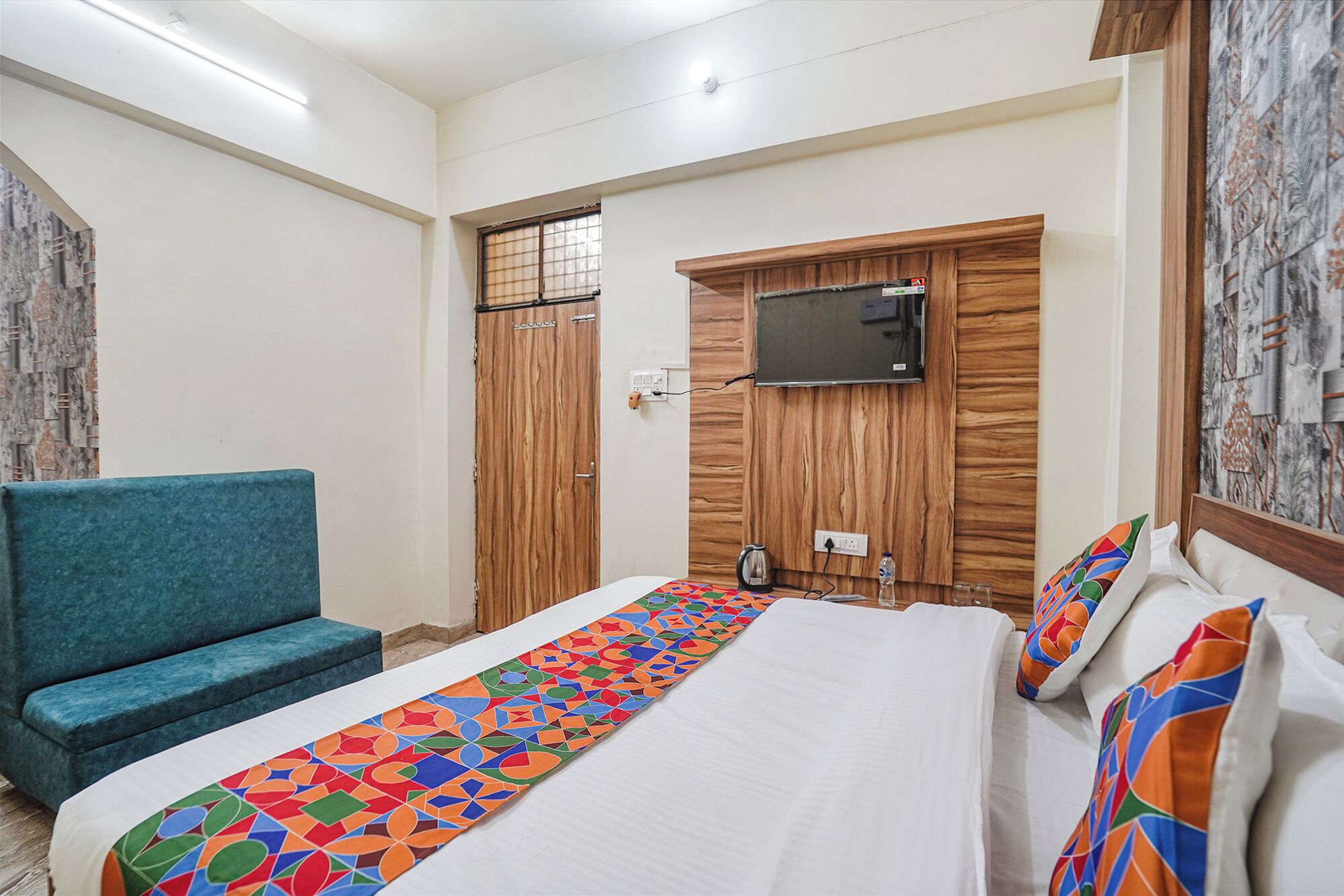 Une chambre d’hôtel avec un lit, un canapé bleu, un téléviseur fixé sur un panneau en bois et un tapis à motifs.