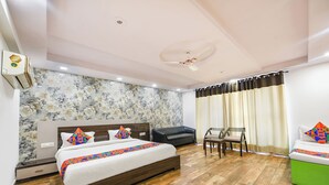 A modern hotel room with a wooden floor, a large bed with colorful pillows, a black leather sofa, a dining table with chairs, and a green ottoman. - Fabhotel Angel Green Residency (Gurugram)