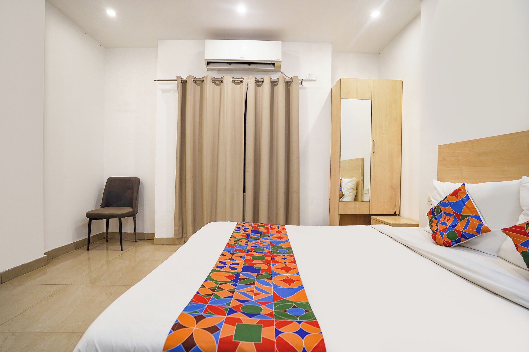 A hotel room with a bed, a chair, a wardrobe, and an air conditioning unit.
