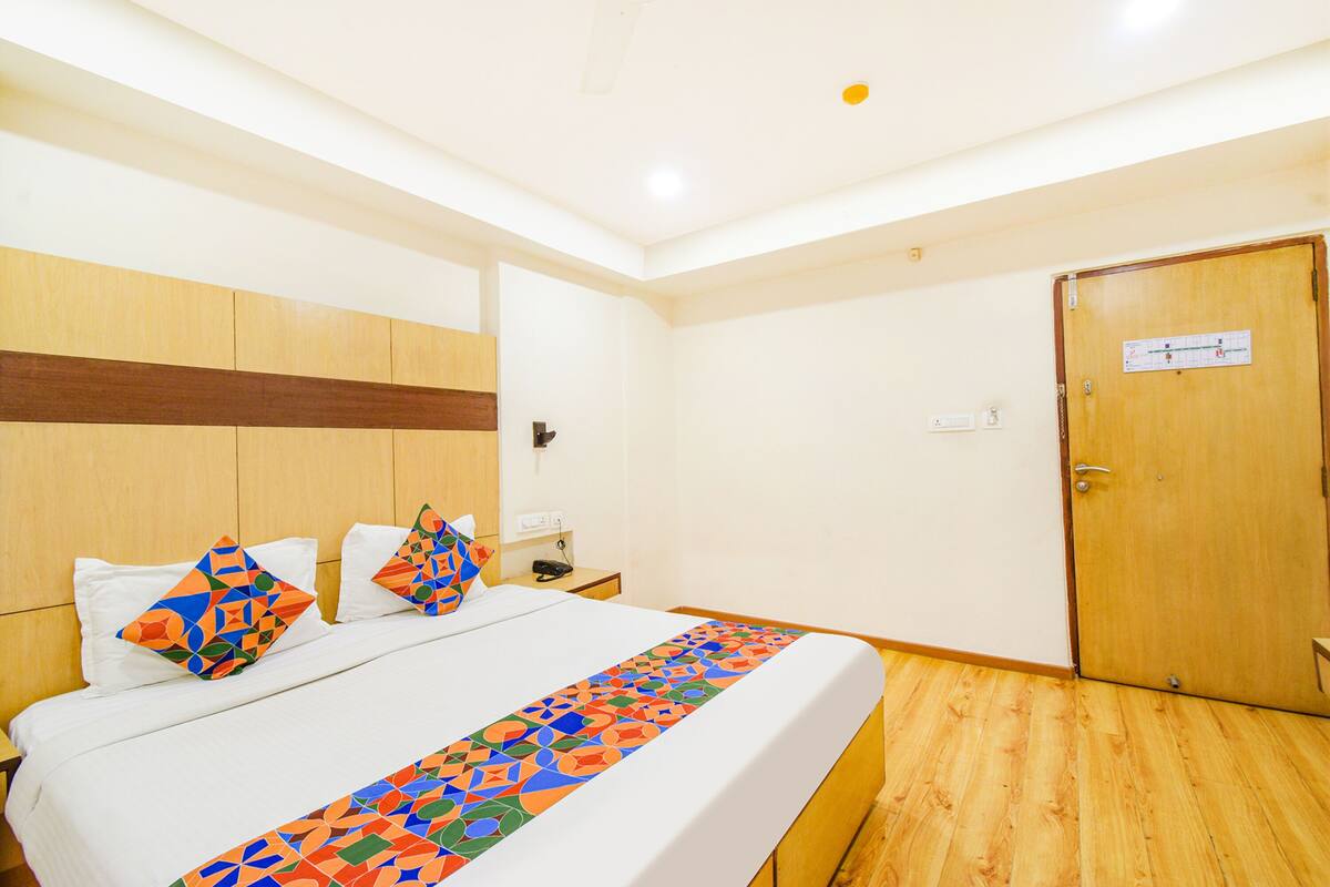 A hotel room with a wooden floor, a bed with a colorful geometric-patterned bedspread, a wooden headboard, a nightstand with a phone and a lamp, and a door with a calendar.