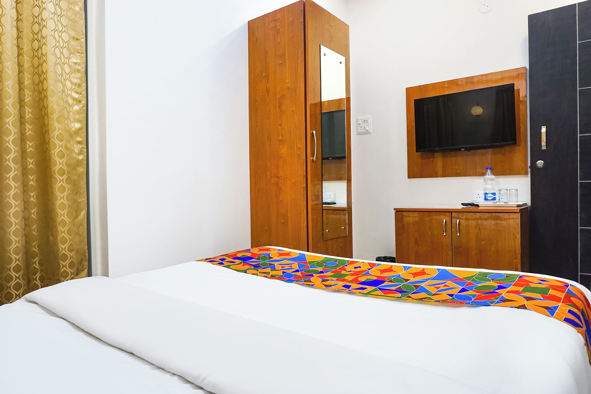 Ein Schlafzimmer mit einem Bett, einem Holzschrank, einem Spiegel, einem Fernseher und einer kleinen Kommode mit einer Wasserflasche und Gläsern.
