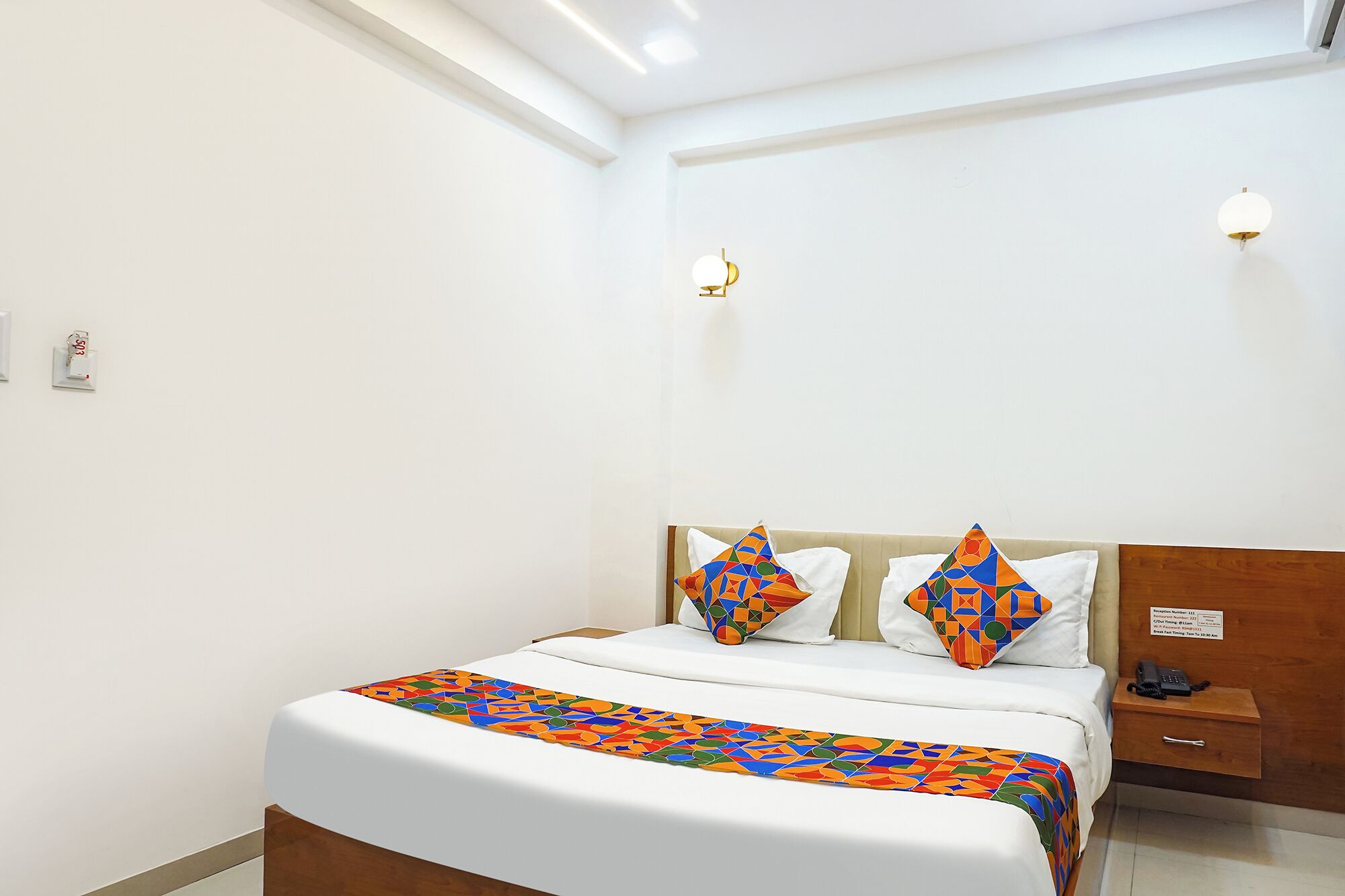 A hotel room with a bed, a bedside table, a nightstand, and a wall-mounted light fixture.
