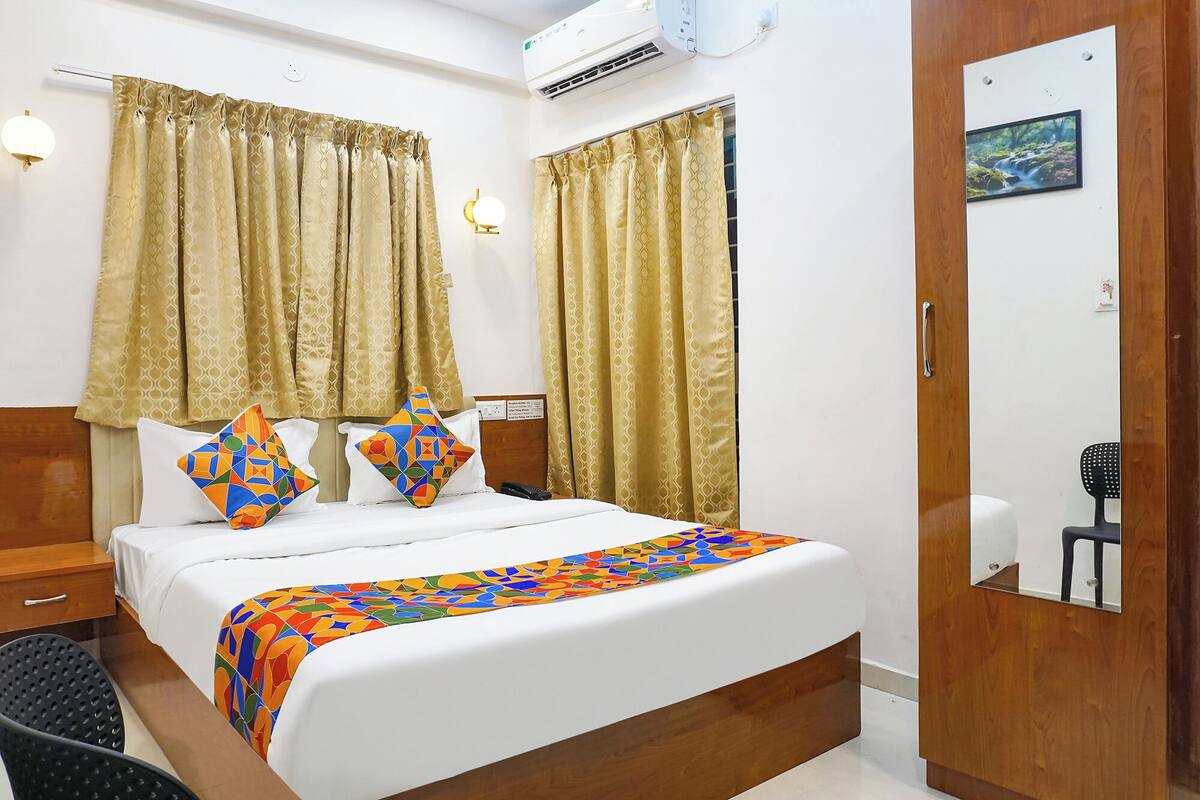 Une chambre d’hôtel avec un lit, deux oreillers, une table de chevet, une chaise, un miroir et une fenêtre avec des rideaux.