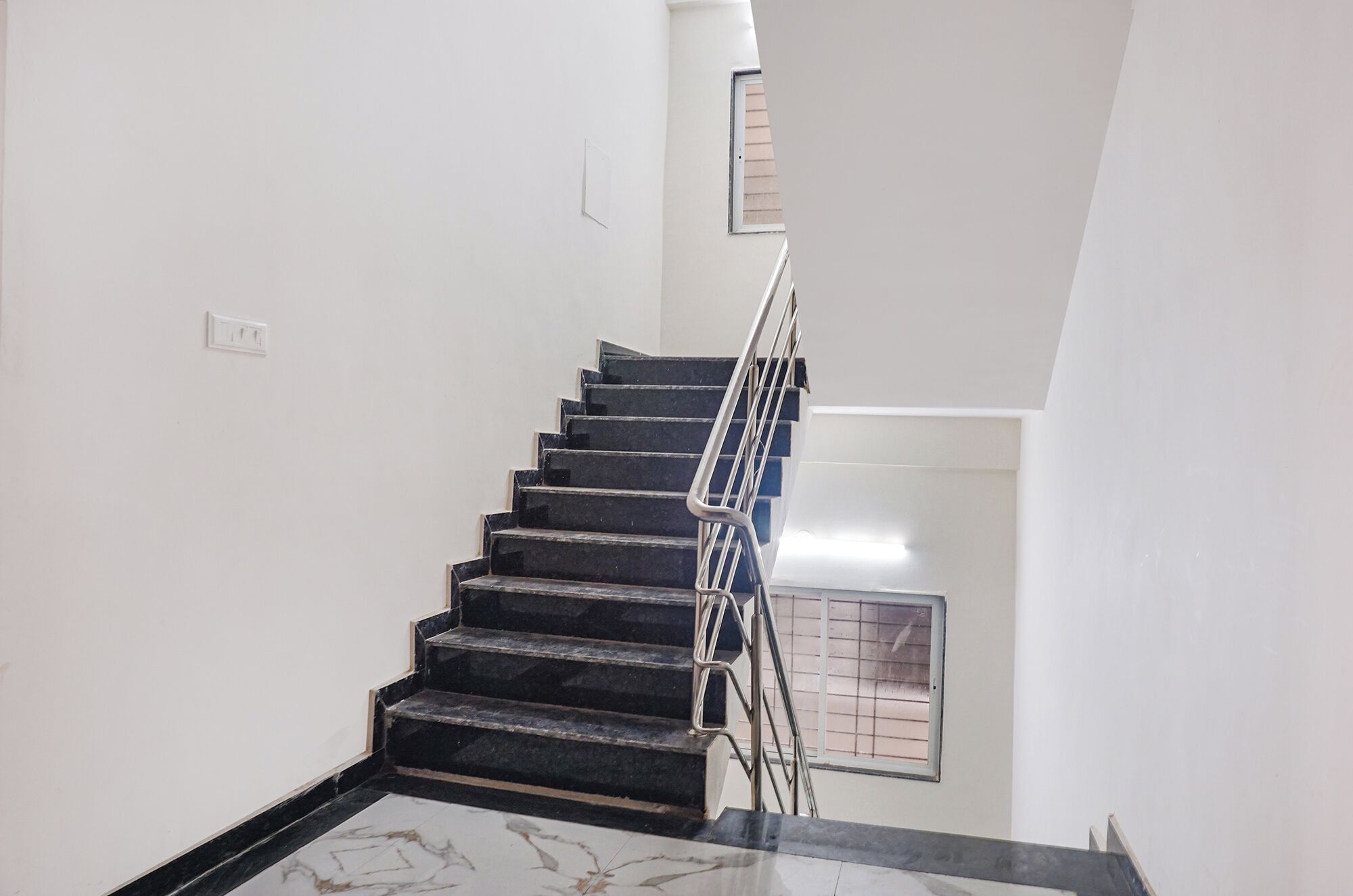 Un escalier avec des marches noires et une rampe métallique, menant à un palier doté d’une fenêtre.