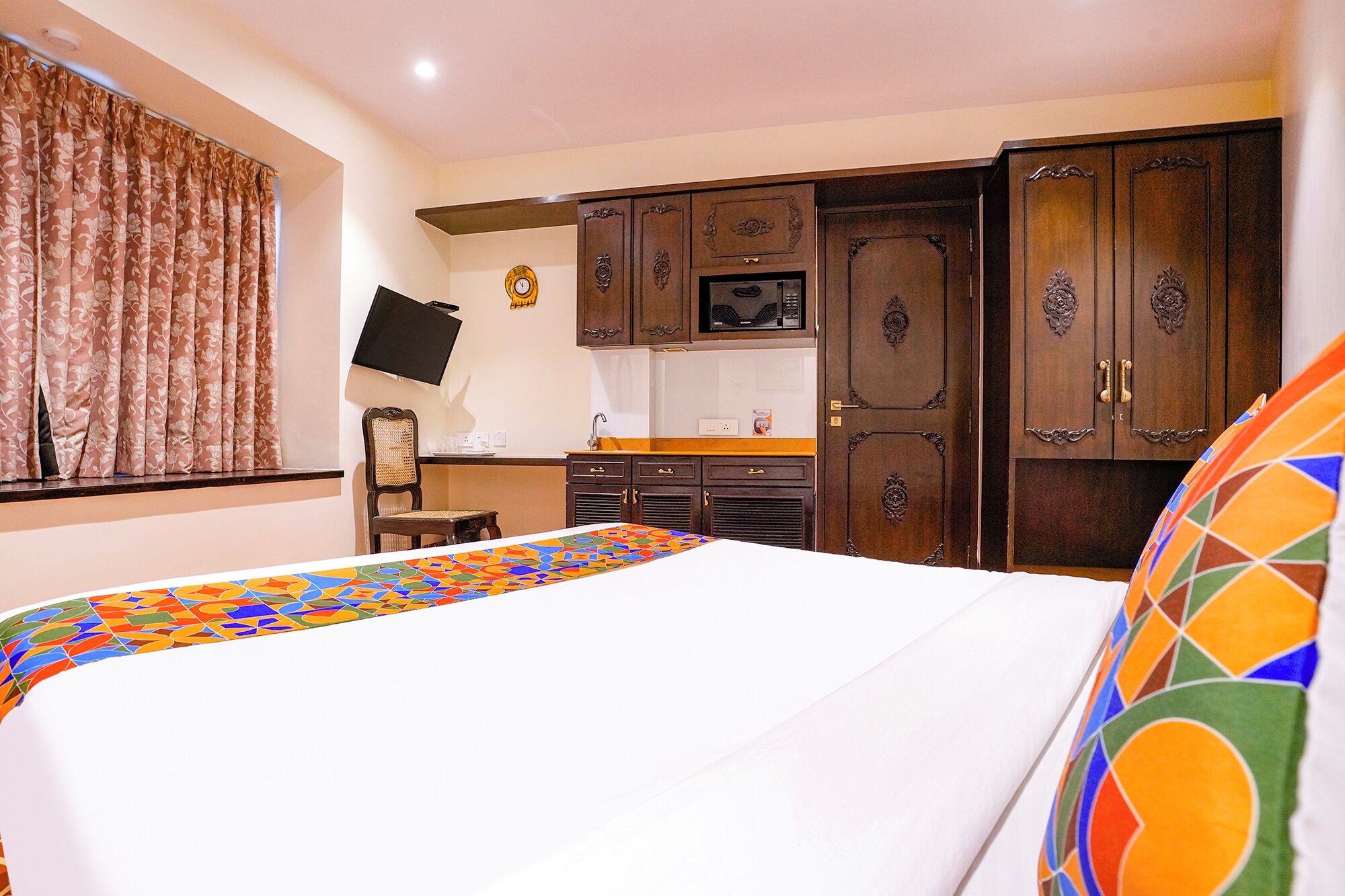 A hotel room with a bed, a TV mounted on the wall, wooden cabinets, and a small kitchen area.