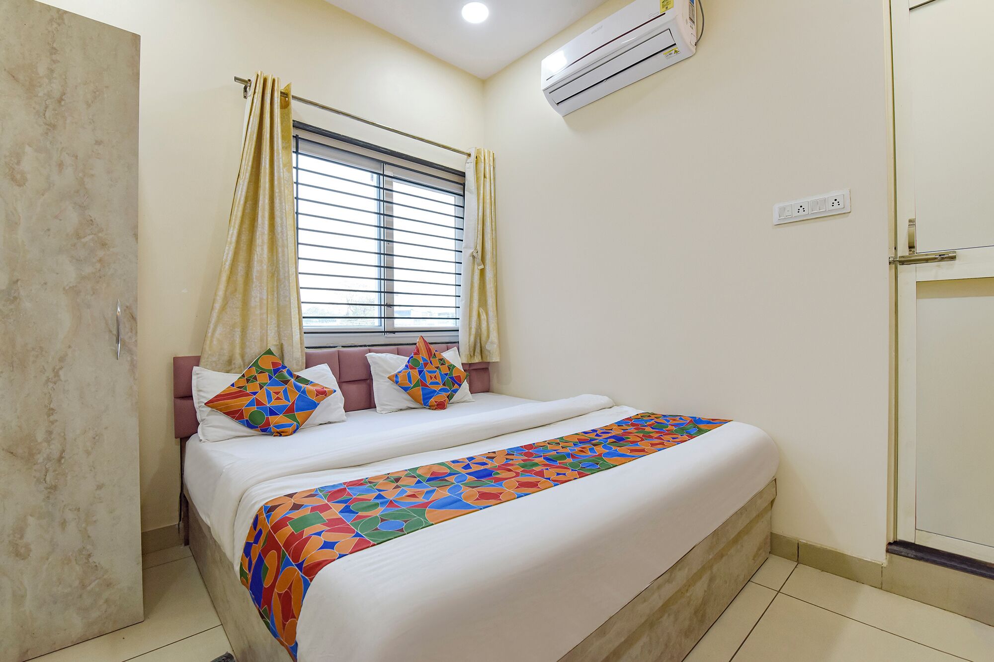 A neatly made double bed with a colorful geometric-patterned bedspread, a window with blinds, and an air conditioning unit on the wall.