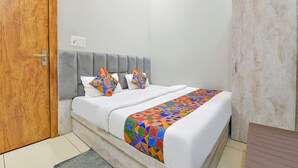 Una camera da letto con una porta in legno, una testiera grigia rivestita in tessuto, un letto bianco con cuscini geometrici colorati e una coperta a motivi.