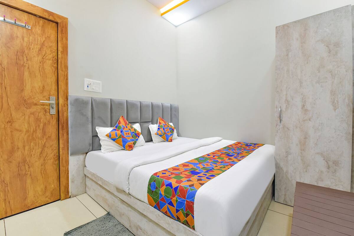 A bedroom with a wooden door, a grey upholstered headboard, a white bed with colorful geometric pillows, and a patterned bedspread.