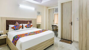 A hotel room with a bed, bedside table, and a desk with a chair. There is a door and an air conditioning unit on the wall.
