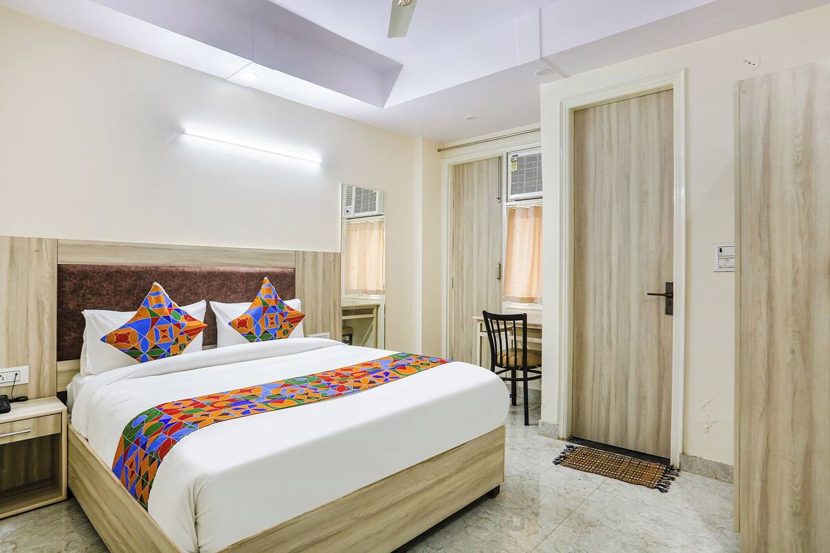 Una camera d'albergo con un letto, un comodino e una scrivania con una sedia. Si vede una porta e un condizionatore d'aria sulla parete.