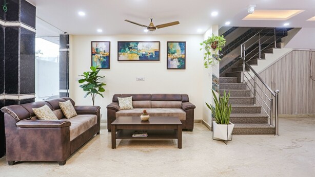 A living room with two sofas, a coffee table, and a ceiling fan. There are three framed artworks on the wall, a staircase, and a potted plant.