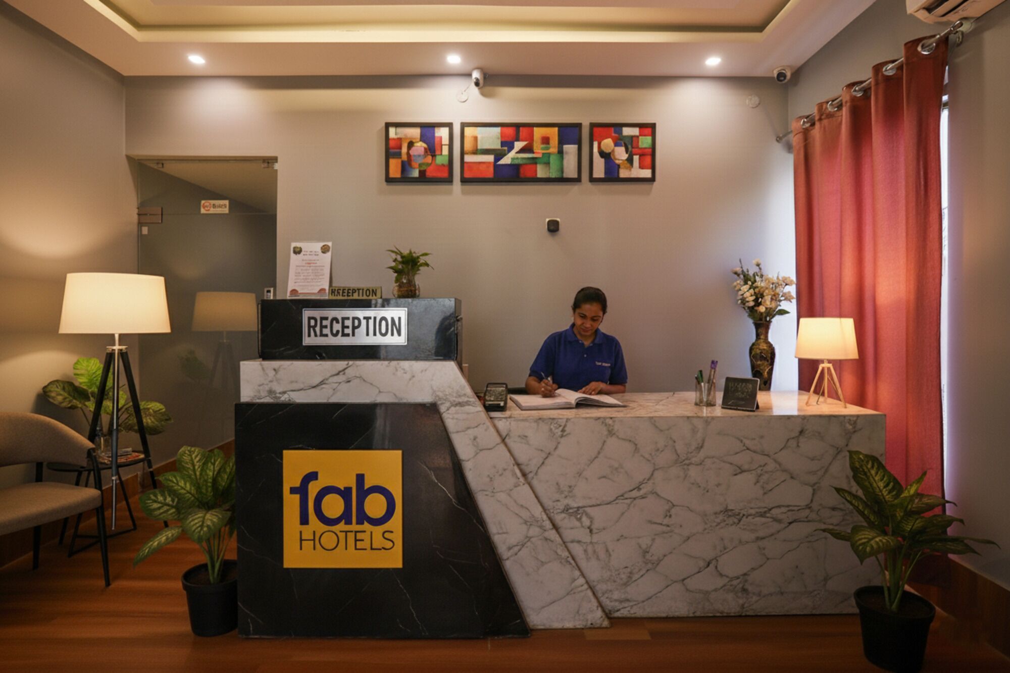 A reception desk with a marble top, a sign reading "Reception", and a "fab hotels" logo. There are two potted plants, a lamp, and a framed abstract painting on the wall.