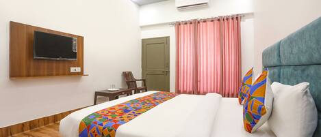 A hotel room with a bed, a TV mounted on the wall, a desk, and a window with curtains.