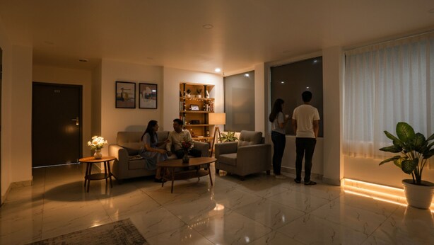 Una sala de estar moderna con sofá, sillones, una mesita de centro y una estantería. Se ve una puerta y una ventana con cortinas.