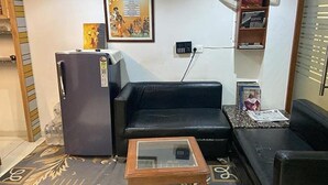 A living room with a black sofa, a glass-top coffee table, a refrigerator, and a wall-mounted TV. - Fabhotel Casa De Grand (Prayagraj)