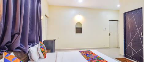 Ein Hotelzimmer mit Bett, Vorhängen, einem Nachttisch, einer Lampe, einer Tür und einem kleinen Fenster.