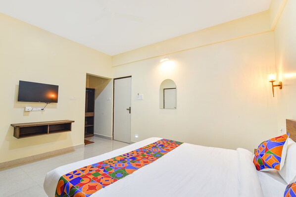 A hotel room with a bed, a TV mounted on the wall, a small shelf, a door, and a wall-mounted light fixture.
