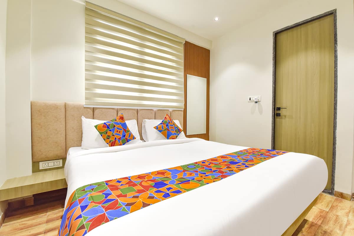 A hotel room with a large bed, a wooden headboard, a window with blinds, a wooden door, and a bedside table with a power outlet.