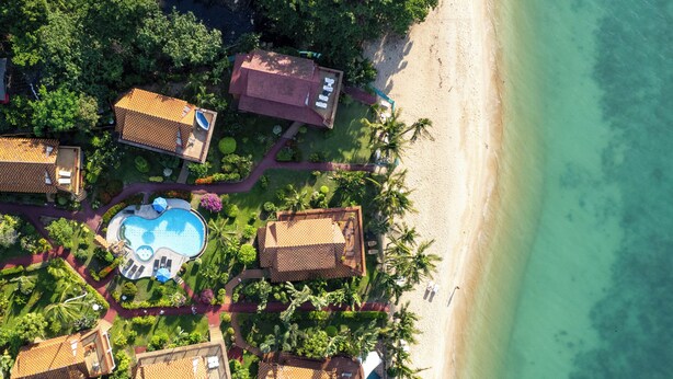 Vista aérea de un complejo turístico con varias edificaciones tipo cabaña, una piscina central y una playa.