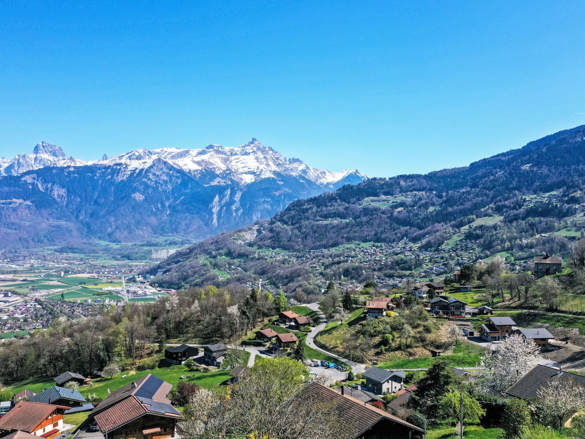 A mountain village with traditional houses, a winding road, and snow-capped mountains in the background.