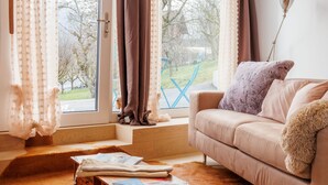 A cozy living room with a beige sofa, a fluffy throw, and a coffee table with magazines. A large window with curtains offers a view of the outdoors. - Lau'tentik by Interhome (Collombey-Muraz)