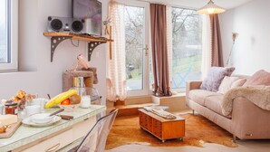 A cozy living room with a sofa, a coffee table, and a television mounted on a wall. There is a window with curtains, a small wooden shelf with a speaker, and a lamp. - Lau'tentik by Interhome (Collombey-Muraz)