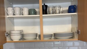 Una cocina con salpicadero de azulejos blancos, un lavamanos de acero inoxidable, una cafetera y estantes con platos y tazas.