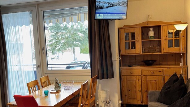 Una habitación con una mesa de comedor de madera, sillas, un sofá gris, un televisor montado en la pared y una zona de cocina con armarios de madera.
