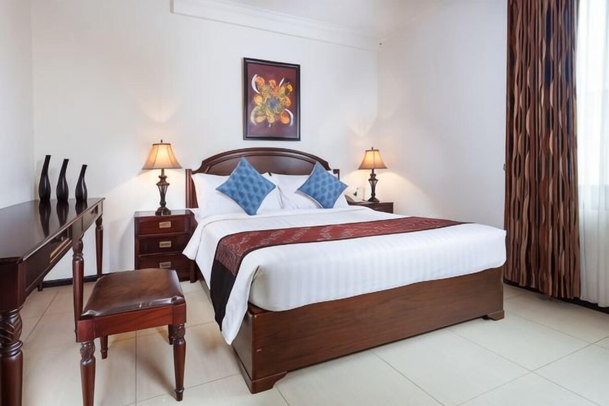 Habitación de hotel con cama de madera, dos lámparas de noche, un escritorio con silla y un adorno en la pared.