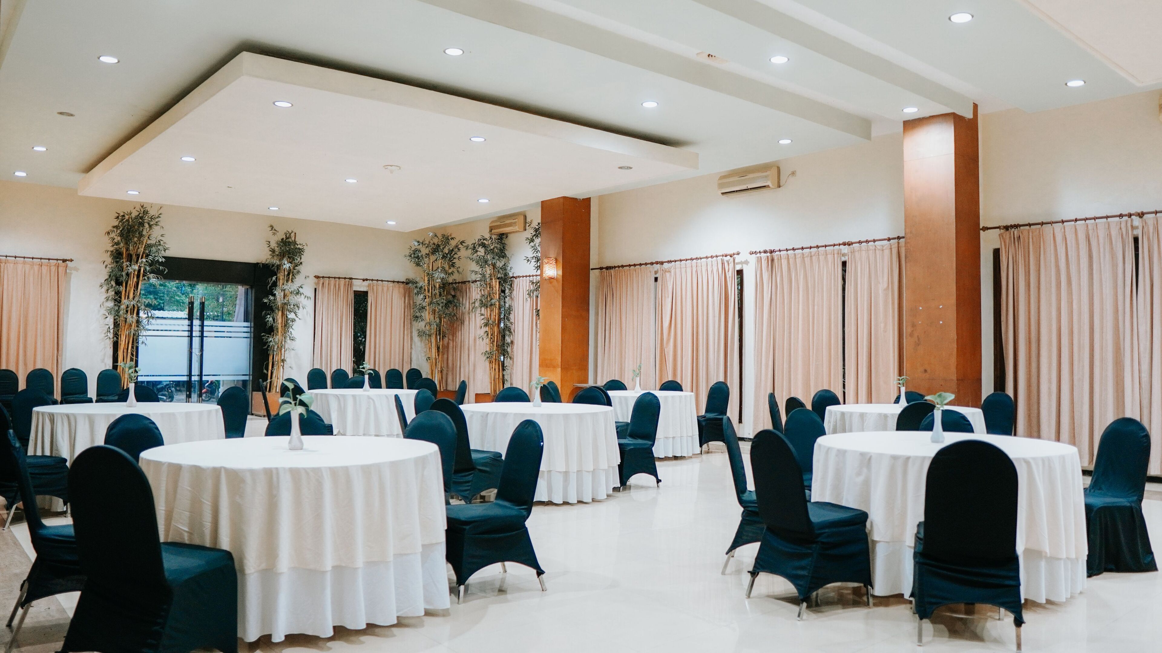 A banquet hall with round tables and chairs, large windows, and a high ceiling with recessed lighting.