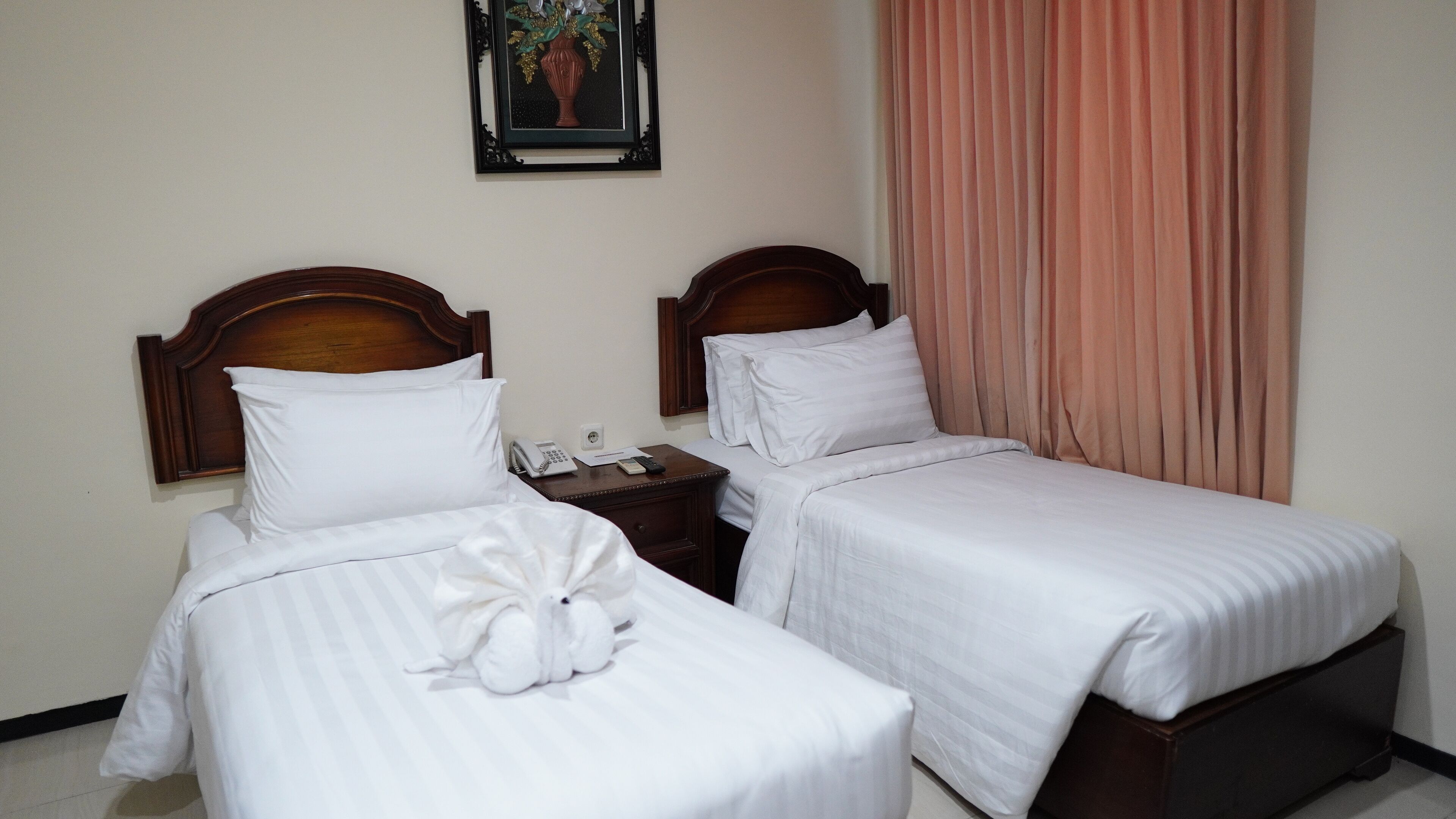 Une chambre d’hôtel avec deux lits simples, une table de chevet avec un téléphone et des documents, et un tableau encadré au mur.