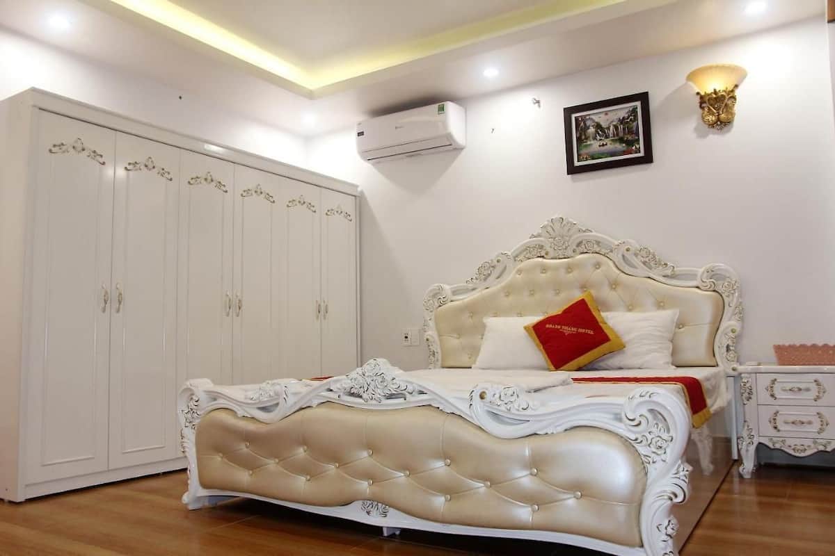 A bedroom with a large white wardrobe, a tufted bed with ornate headboard, and a bedside table with a lamp.