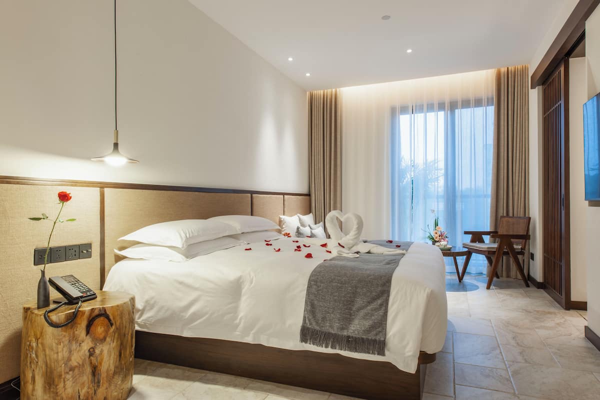 Une chambre d’hôtel avec un grand lit, une table de chevet en bois ornée d’un vase et d’un téléphone, un bureau avec une chaise, et une grande fenêtre avec des rideaux.