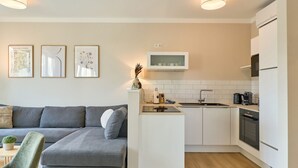 Et moderne køkken med hvide skabe, en indbygget ovn og en mikrobølgeovn. Der er en grå sofa med et lille bord og en grøn stol i opholdsstuen.