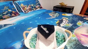 Un letto con una coperta a tema oceano blu, un cesto bianco pieno di carta sfilacciata e un asciugamano nero con la scritta "Makeup".